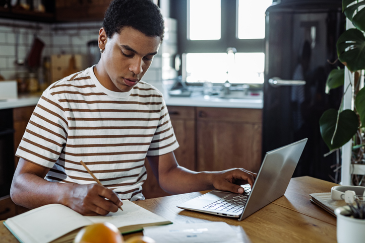 Young man writing notes while using laptop