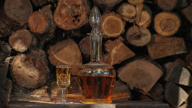 Cognac pouring in the glass from bottle. Hand puts a carafe with golden cognac and pouring into the glass, wooden table near logs. Strong drink - brandy or whiskey close-up. Alcohol amber drink