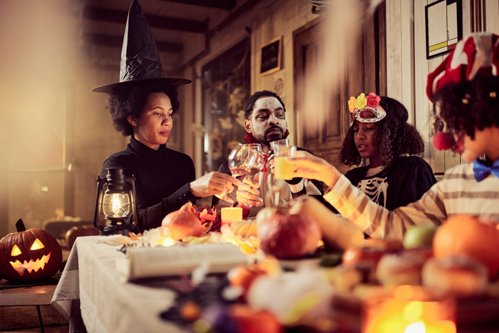 Black family in costumes toasting on Halloween at dining table.