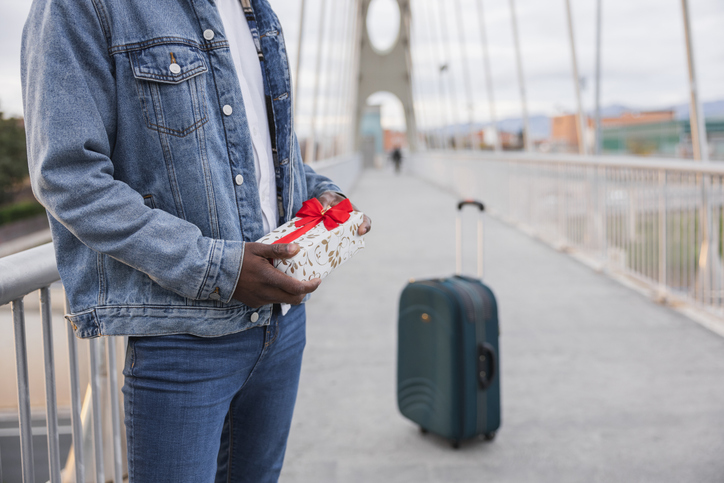 Young man holding a gift after returning from trip, standing on a bridge with his luggage.