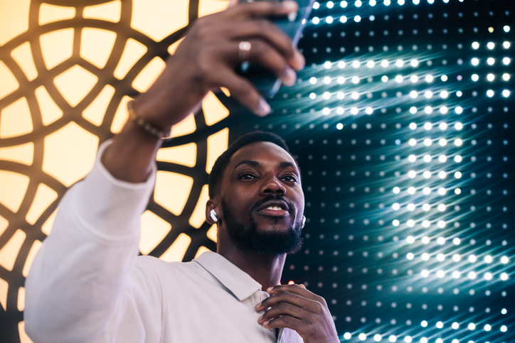 A close-up of a joyful man taking a selfie in front of a colorful LED wall, his face glowing with excitement as he lifts his phone.
